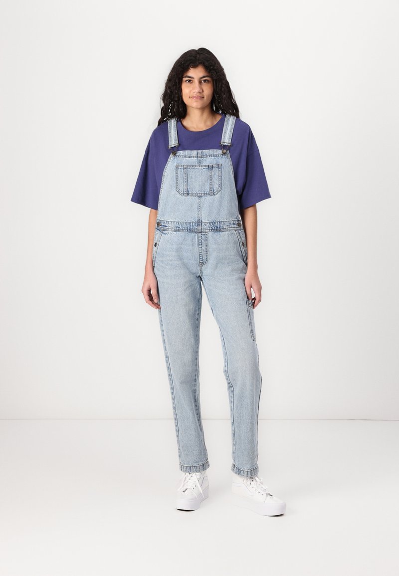 Salopette en denim bleu clair avec une coupe ample, grande poche avant et bretelles ajustables, associée à un t-shirt violet et des baskets blanches.