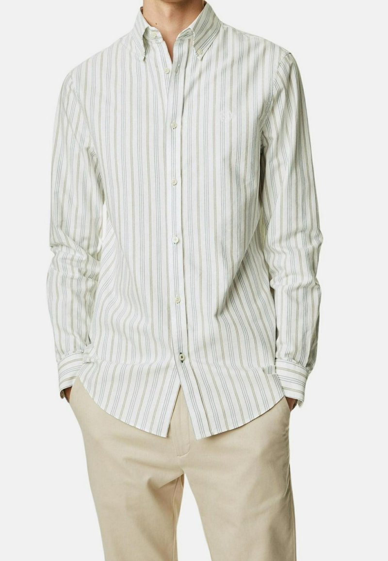 Camisa blanca de botones con rayas verticales verdes, de mangas largas y con un logo en el pecho izquierdo; combinada con pantalones beige claros.