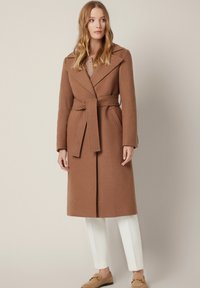 Brązowy, wełniany płaszcz z paskiem, kołnierzem z wycięciem i bocznymi kieszeniami, połączony z białymi spodniami i beżowymi mokasynami. Gładka faktura, dopasowany krój.