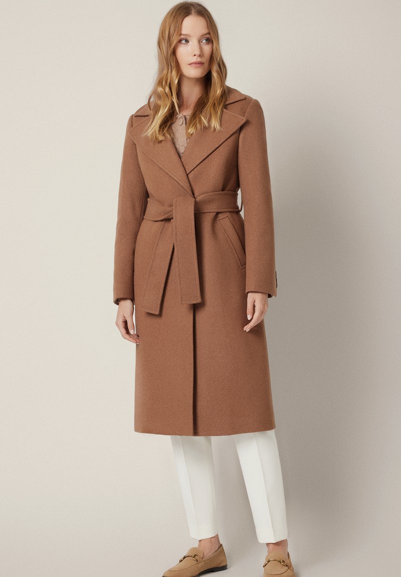 Brązowy, wełniany płaszcz z paskiem, kołnierzem z wycięciem i bocznymi kieszeniami, połączony z białymi spodniami i beżowymi mokasynami. Gładka faktura, dopasowany krój.