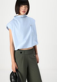 Top corto azzurro chiaro con colletto alto, maniche corte e una spilla decorativa argentata, abbinato a pantaloni verdi oliva a vita alta.