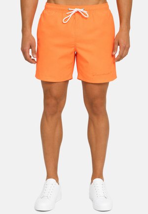 Homme portant un short de bain orange vif avec cordon de serrage et des baskets blanches, debout, les bras détendus le long du corps, sur un fond uni.