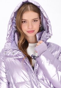 Irideszcens lila puffermellény kapucnival, patentgombokkal, bordázott mandzsettákkal és steppelt mintával. Belső részén lágy bélés látható.