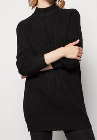 Femme portant une robe pull longue noire avec un col haut, un motif texturé et des poignets côtelés, posant avec les bras croisés.