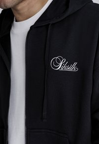 Čierna mikina na zips vyrobená z mäkkého materiálu, s bielym nápisom na ľavom prsníku, s kapucňou na šnúrku a prednými vreckami.