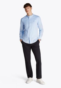 Lyseblå skjorte med knappelukking og ståkrage, brukt sammen med mørke bukser og hvite joggesko. Glatt stoff, minimalistisk design og diskret logodetalj.