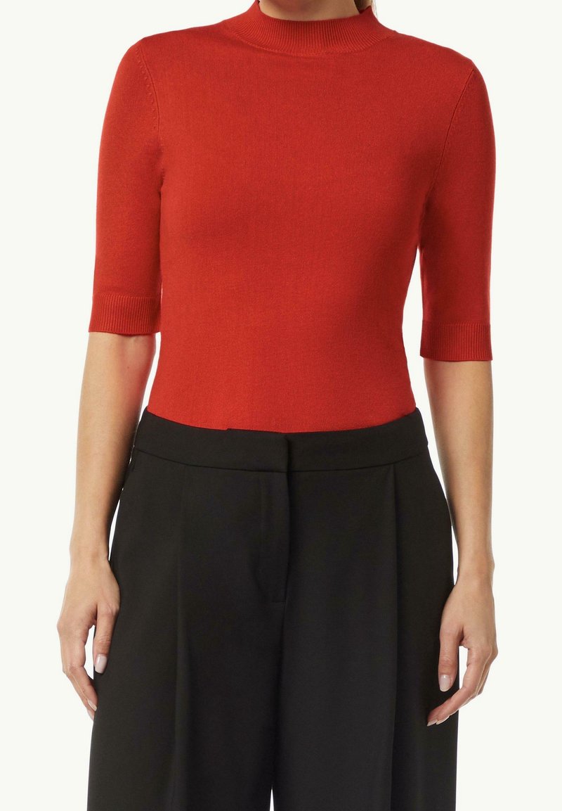 Top de punto acanalado rojo con cuello alto y mangas cortas, combinado con pantalones negros a medida. Textura suave y diseño ajustado.