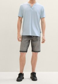 Camisa henley de manga corta azul claro combinada con pantalones cortos grises y zapatillas negras, modelada por un hombre de pie frente a un fondo neutro.
