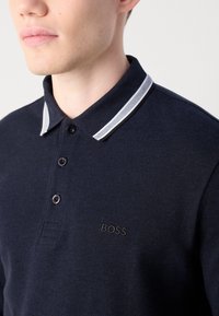 Vyras vilkintis tamsiai mėlyną polo marškinėlį su baltais ir juodais dryžiais ant apykaklės ir išsiuvinėtu "BOSS" logotipu ant krūtinės.