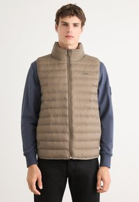 Beige puffer-vest met verticale naden, hoge kraag en ritssluiting aan de voorkant. Draag het over een blauwe lange mouwen shirt, met een logo op de vest.