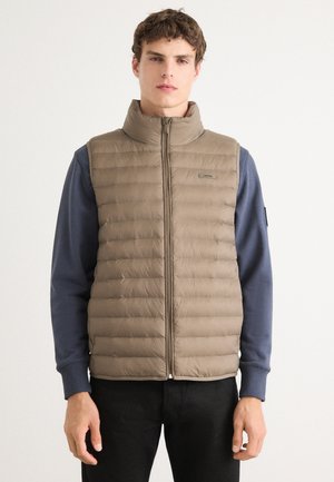 Beige puffer-vest met verticale naden, hoge kraag en ritssluiting aan de voorkant. Draag het over een blauwe lange mouwen shirt, met een logo op de vest.