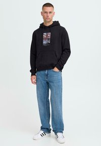Joven de pie con la mano en el bolsillo, vistiendo una sudadera negra con estampado gráfico, vaqueros azules y zapatillas blancas con franjas negras.