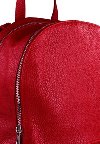 Sac à dos en cuir rouge avec une surface texturée, une forme arrondie et un détail de fermeture éclair argenté sur le côté.