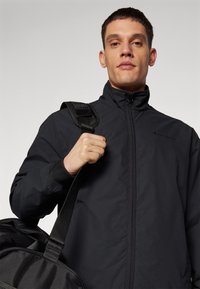 Chaqueta negra con cremallera y cuello alto, con puños elásticos. El portador sostiene un bolso de viaje negro con una correa texturizada.