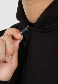 Felpa nera con zip frontale, realizzata in tessuto morbido. Presenta un cappuccio e cuciture a vista. Una mano sta parzialmente aprendo il colletto.