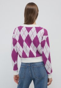 Femme portant un pull en argyle violet et blanc à manches longues et un jean en denim bleu, se tenant contre un fond uni.