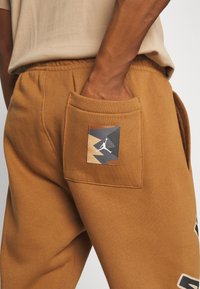 Person klädd i bruna sweatpants med en fickpatch som visar en geometrisk design och en vit siluett som hoppar med en basketboll.