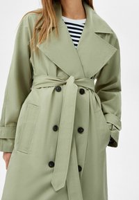 Jasnozielony trencz z podwójnym zapięciem, materiałowym paskiem i dwoma przednimi kieszeniami; wyróżnia się dużymi czarnymi guzikami i szerokimi klapami.