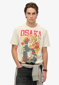 Camiseta de algodón color crema con un gráfico de tigre colorido, la palabra "OSAKA" en rojo y detalles en gris. Textura suave, diseño de cuello redondo.