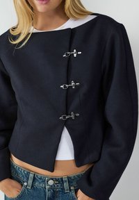 Veste cropped navy à manches longues, présentant des fermetures à anneaux en argent et un design épuré et minimaliste. Portée par-dessus un top blanc et un jean.