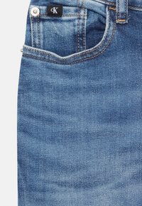 Jeans de mezclilla azul con un bolsillo delantero que tiene una etiqueta, lavado claro, costuras naranjas y herrajes plateados en la cintura.