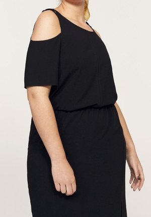 Kvinde iført en sort kjole med bare skuldre og elastisk taljebånd, stående mod en ensfarvet lys baggrund.