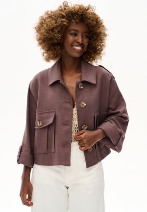 Uśmiechnięta kobieta z lokowanymi włosami, ubrana w brązową zapinaną na guziki kurtkę, beżowy sweter i białe spodnie, na jednolitym tle.