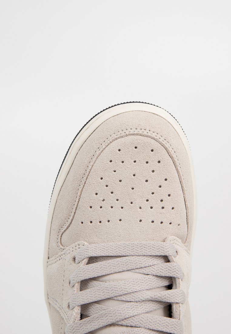Zapatilla de gamuza beige clara con área del dedo perforada, cierre de cordones plano y suela de goma negra contrastante. Textura suave y diseño sencillo.