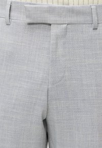 Gros plan d'un pantalon en tissu gris tissé montrant des passants de ceinture, une ceinture et un détail de braguette avant.