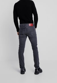 Jeans de mezclilla gris con un corte ajustado, que incluyen un parche de cuero rojo en la parte posterior de la cintura y dos bolsillos traseros. Zapatos negros visibles.
