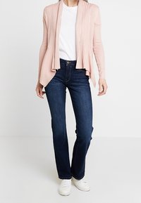 Frau trägt eine hellrosa offene Strickjacke über einem weißen T-Shirt, dunkle Bootcut-Jeans und weiße Sneaker, steht vor einem weißen Hintergrund.