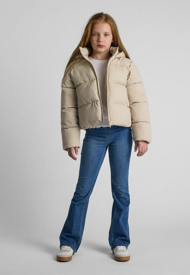 Giubbotto puffa beige con zip frontale, design voluminoso, colletto alto, indossato sopra un top chiaro, abbinato a jeans a zampa d'elefante blu e sneakers bianche.