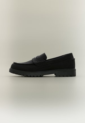 Mocassin en cuir noir avec une semelle rainurée, coutures moc toe et un détail de bride sur le dessus, vu de profil sur un fond neutre.