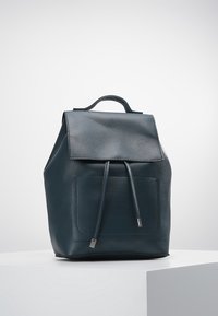 Ciemnozielona skórzana plecak z klapą, detalami w postaci sznurków, zewnętrzną kieszenią z przodu i mocnym uchwytem na górze. Gładka tekstura.