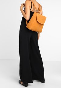 Mochila de ante color tostado con acabado suave, cierre de solapa redondeada, correas ajustables y diseño minimalista. Presentada sobre un fondo liso.