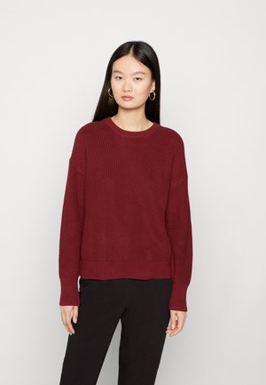Burgunderfarbener Strickpullover mit rundem Ausschnitt, gerippter Struktur, überschnittenen Schultern und einem leicht verkürzten Saum. Kombiniert mit schwarzen Hosen.