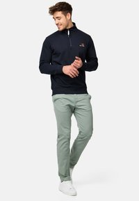 Pulóver de media cremallera de color marino en tejido suave con puños y dobladillo acanalados, combinado con chinos verde claro y zapatillas blancas. Detalle de logo bordado.