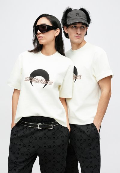 White cotton t-shirts with logo, black patterned pants, silver hardware belt, black sunglasses, and a black cap with faux fur accents.