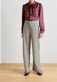 Blusa di raso bordeaux con fiocco, maniche lunghe; pantaloni a palazzo a quadri a vita alta; scarpe nere lucide con dettagli e fibbie.