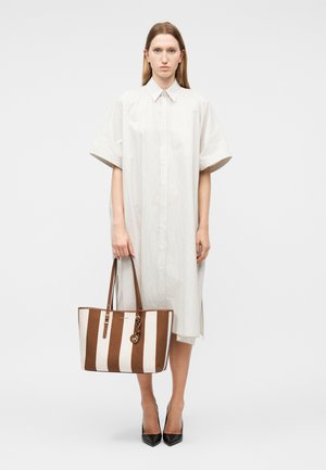 Γυναίκα με λευκό, φαρδύ φόρεμα με κοντά μανίκια, κρατώντας μια μεγάλη καφέ και λευκή τσάντα ώμου με ρίγες, φοράει μαύρες μυτερές γόβες, στέκεται σε λευκό φόντο.