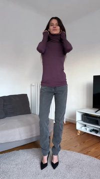 Long-sleeve purple turtleneck, fitted design, paired with high-waisted gray jeans and black pointed heels, standing on a textured rug.