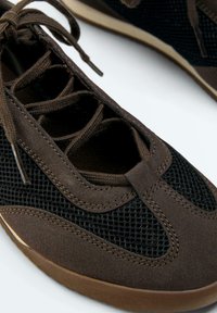 Gros plan d'une basket décontractée marron et noire avec tissu en mesh, détails en daim et lacets marron sur fond blanc.