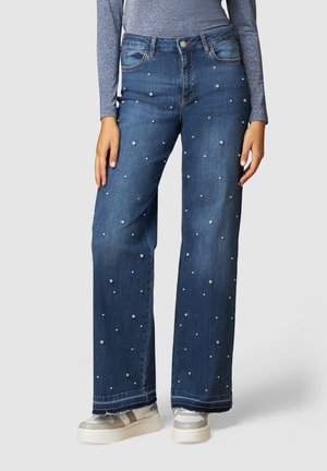 Femme portant un jean bleu large décoré de petites perles éparpillées, associé à des baskets grises et un haut gris à manches longues.