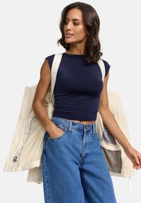 Top court en marine à manches courtes, associé à un jean en denim léger bleu clair taille haute. Veste beige portée sur les épaules montrant un intérieur à motifs.
