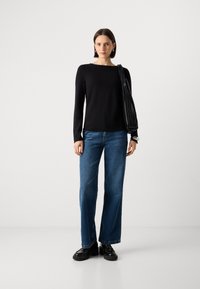 Schwarzes Langarmoberteil mit eckigem Ausschnitt, kombiniert mit hoch taillierten blauen Jeans und klobigen schwarzen Schuhen; Frau hält eine schwarze plissierte Tasche.