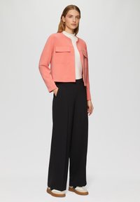 Korallfarbene cropped Jacke mit zwei Fronttaschen, kombiniert mit schwarzen weit geschnittenen Hosen und cremefarbenen Sneakers. Glatte Textur, lässige Passform und modernes Design.