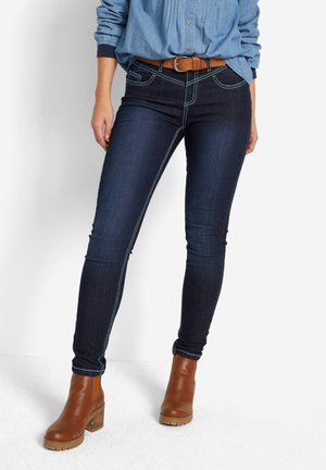 Jeans skinny bleu marine en denim, avec des coutures contrastées, cinq poches et une taille mi-haute. Associés à des bottes chevilles marron.
