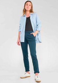 Chemise boutonnée rayée bleue et blanche, débardeur noir, pantalon teal, et baskets blanches avec semelles crème, posture debout, mains dans les poches.