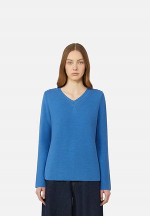 Giovane donna con capelli castani lisci indossa un maglione blu a collo a V e maniche lunghe e pantaloni scuri, in piedi davanti a uno sfondo bianco.