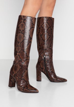 Bottes marron montant jusqu'aux genoux avec motif écaille de serpent, bouts pointus, talons carrés et fermetures éclair latérales portées sur des jambes nues.